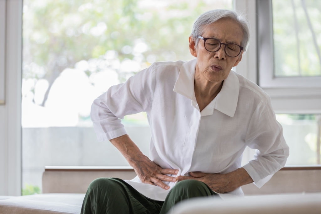 Sick senior patient sitting on a bed, holding her lower abdomen and showing signs of pain or discomfort.