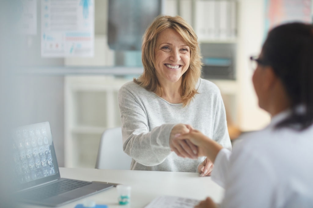 Mature happy patient sitting at the table and greeting with her doctor and smiling to her during her visit at clinic