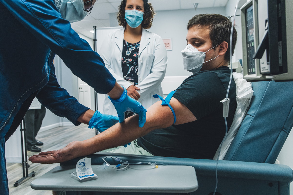 A healthcare worker prepares a patient for a blood draw while a doctor observes, all wearing masks in a clinical setting.