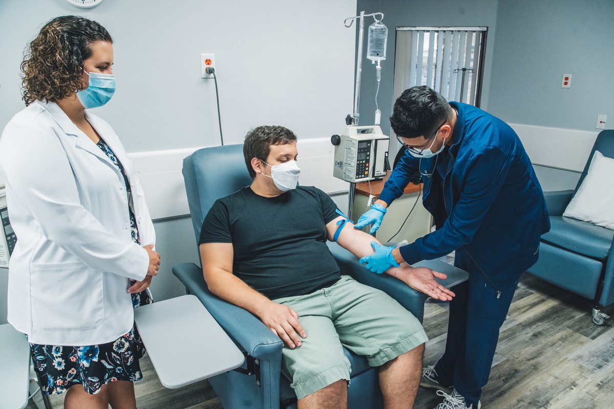 A healthcare worker prepares a patient for an IV procedure while a medical professional observes in a clinical treatment room.