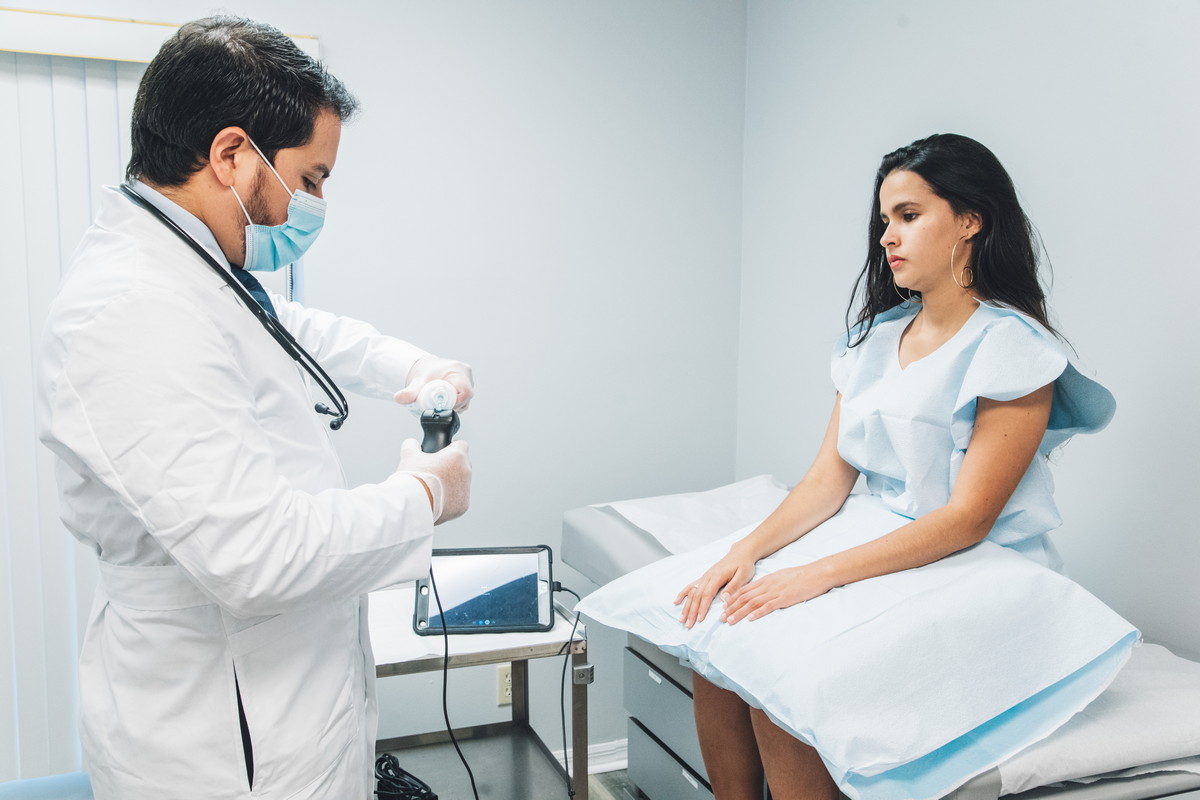 A doctor prepares an instrument while a patient sits on an exam table wearing a medical gown.
