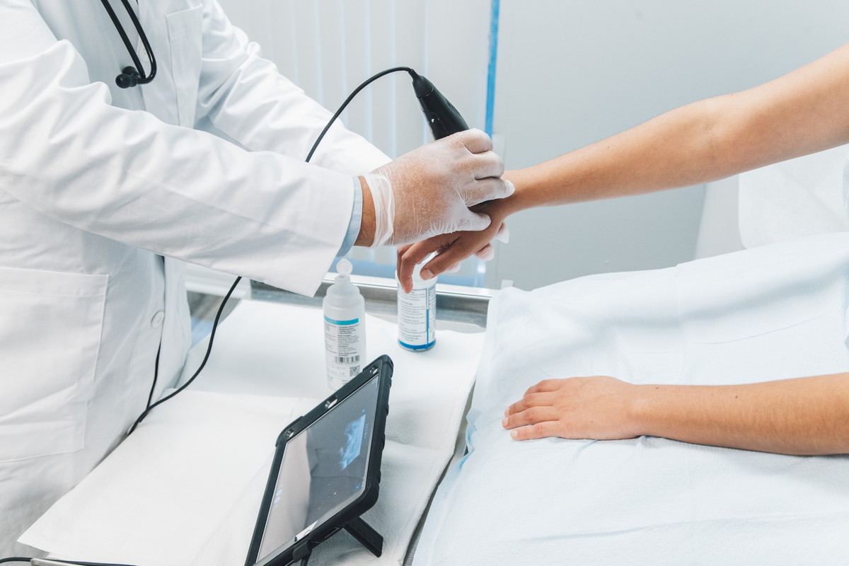 A doctor performs an ultrasound scan on a patient's arm using a handheld device, with the image displayed on a nearby tablet.