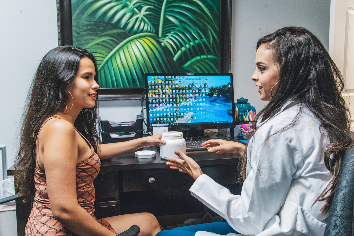 A doctor consults with a patient in an office, discussing a skincare product while seated at a desk with a computer.